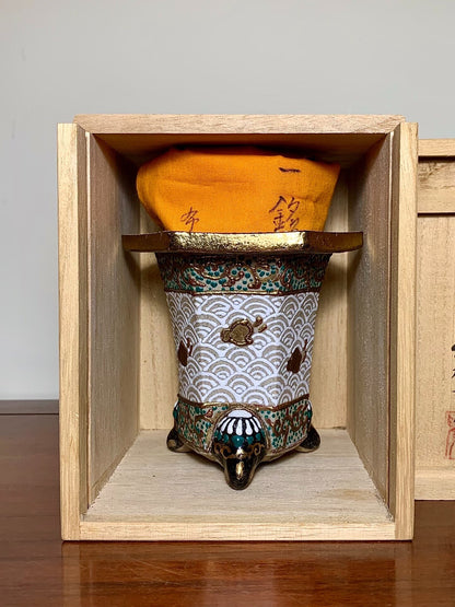 A small, three-legged ceramic pot with wave and turtle patterns, glazed in white, green, and gold, sits in a wooden box.