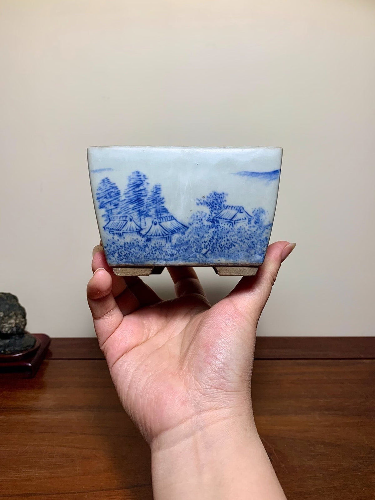 A hand holds a small, white, square ceramic pot with blue landscape painting.