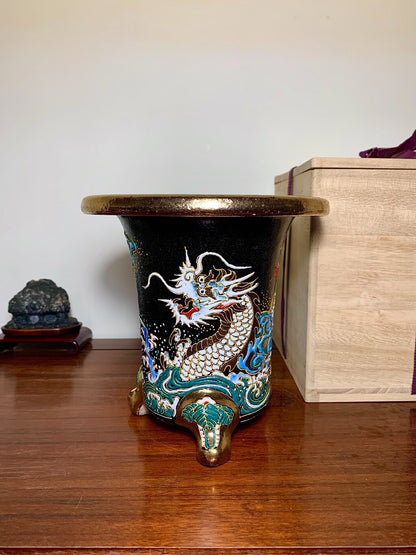 Black glazed pot with a white and gold dragon on a blue wave, three bronze feet.
