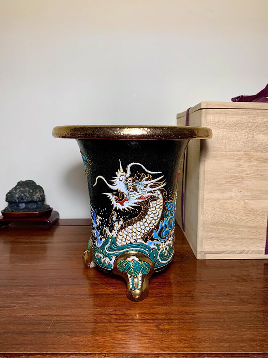 Black glazed pot with a white and gold dragon on a blue wave, three bronze feet.