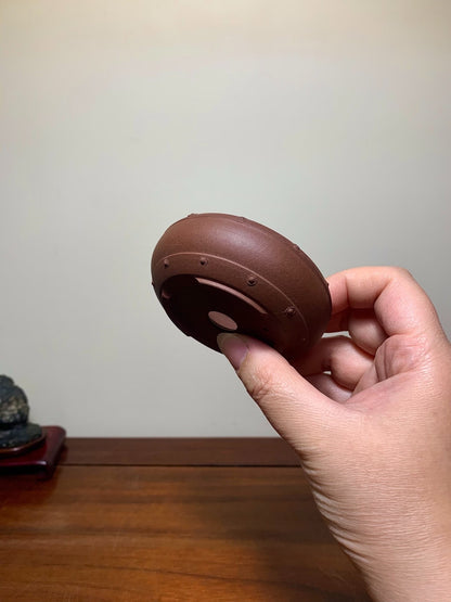 A hand holds a brown, unglazed, round bonsai pot with small raised dots and a central circular mark.