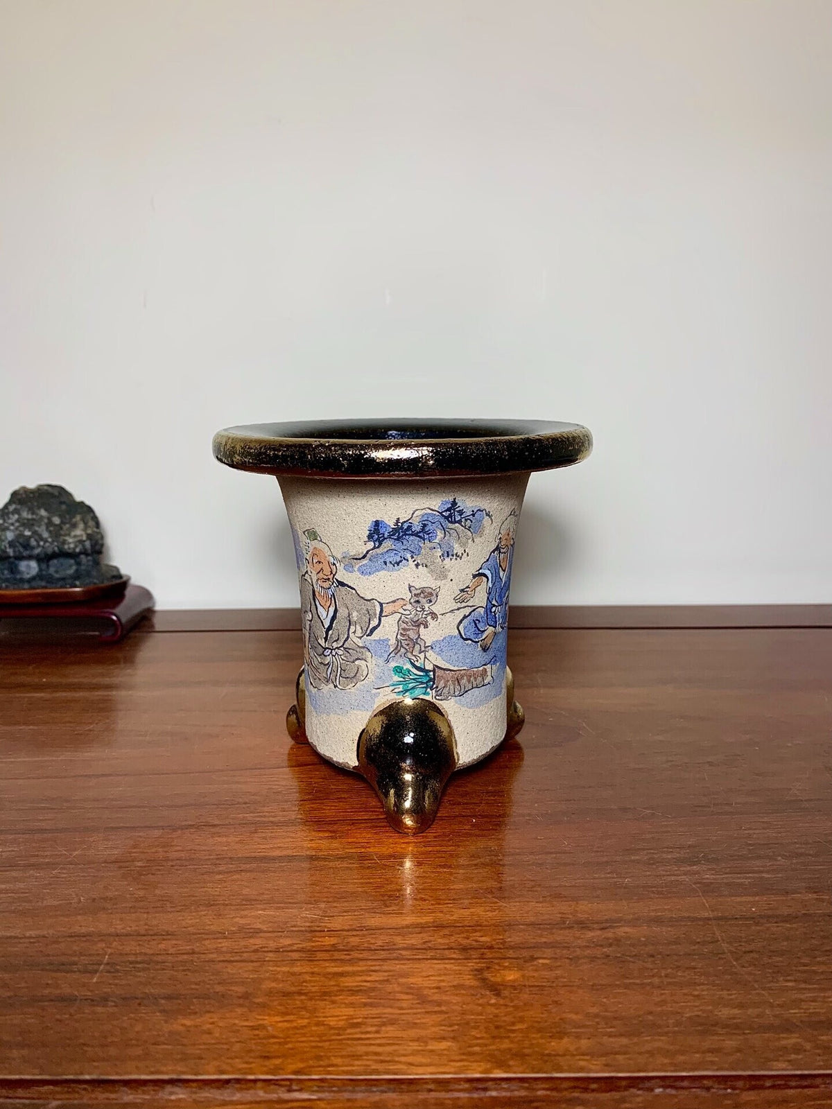 A beige ceramic pot with blue painted figures, black rim, and three bronze feet.