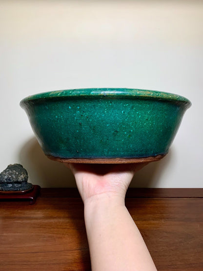 A hand holds a glossy, deep green glazed ceramic pot with a wide rim and unglazed brown base.