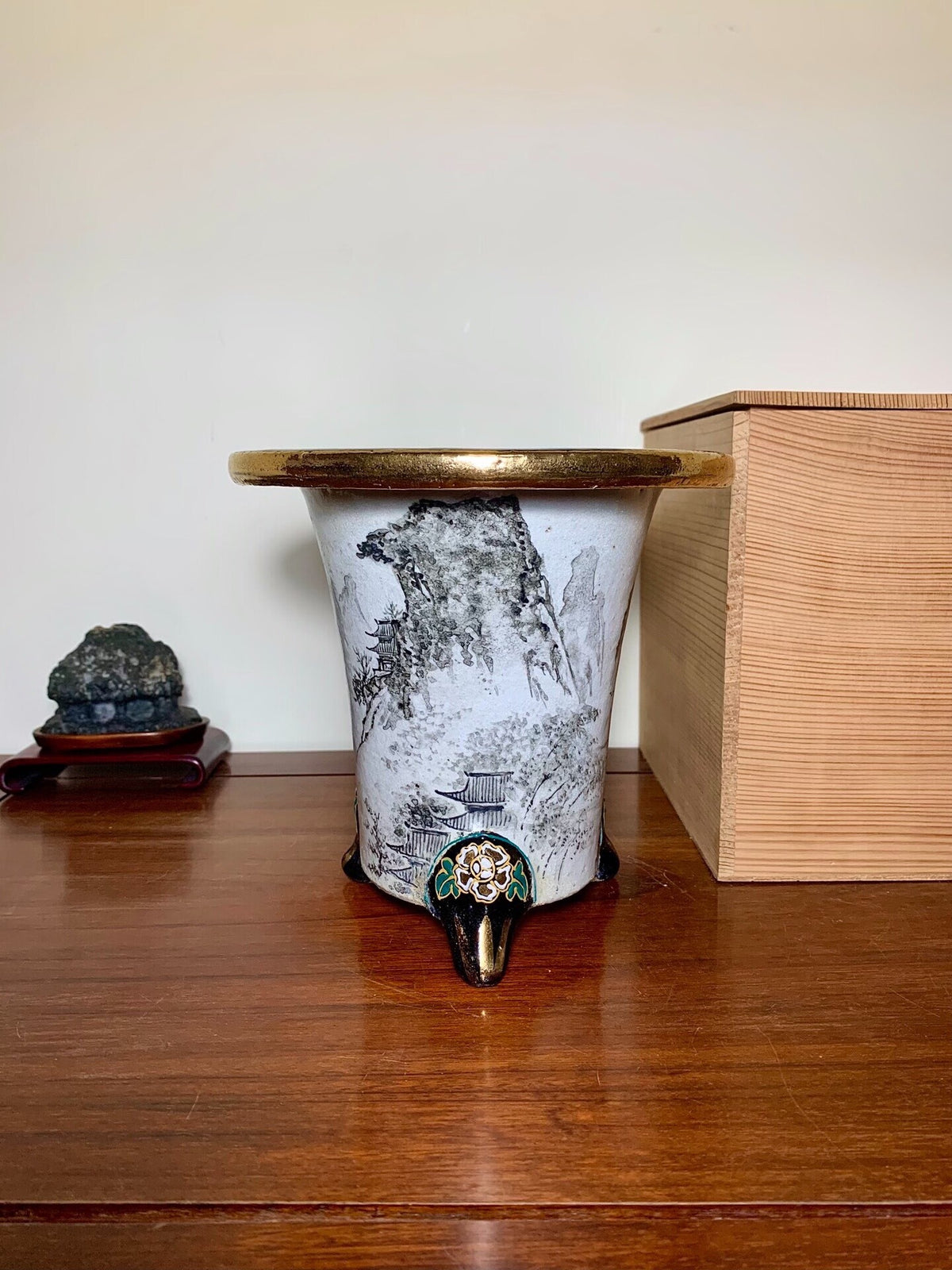 White glazed pot with black mountain scene and gold rim, three legs with ornate metal base.