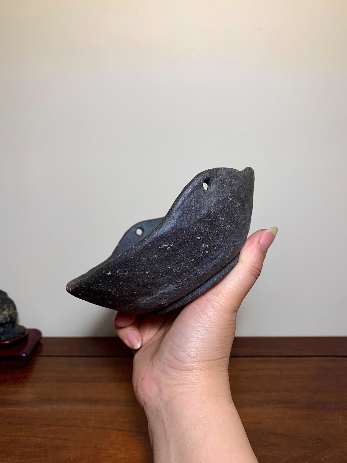 A hand holds a dark, unglazed ceramic pot with a curved rim and two small holes.