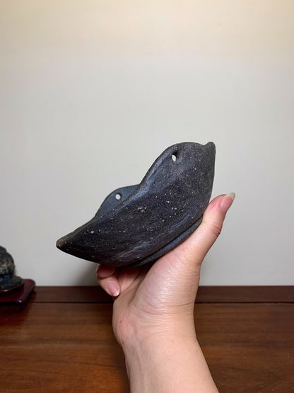 A hand holds a dark, unglazed ceramic pot with a curved rim and two small holes.