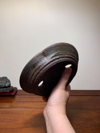 A hand holds a dark brown, glazed, oval-shaped pot with two holes and concentric ridges.