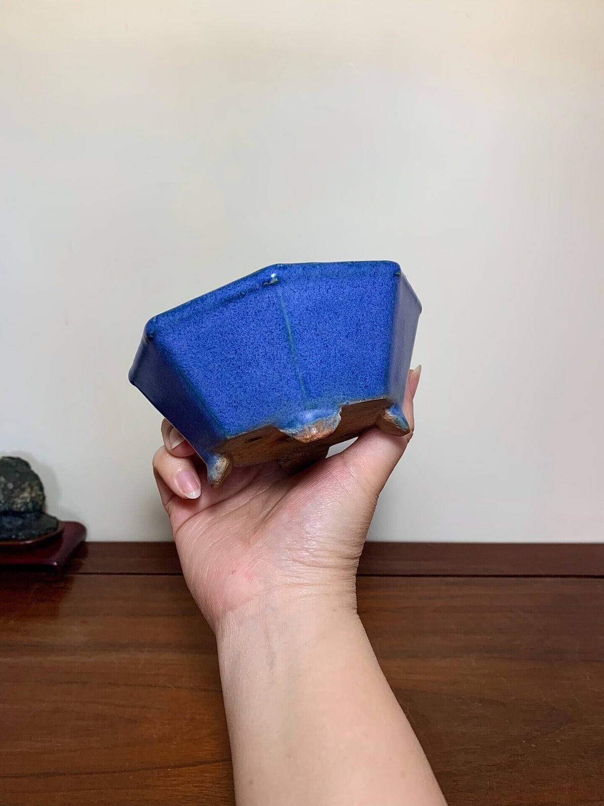 A hand holds a hexagonal, glossy blue glazed pot with unglazed brown feet.