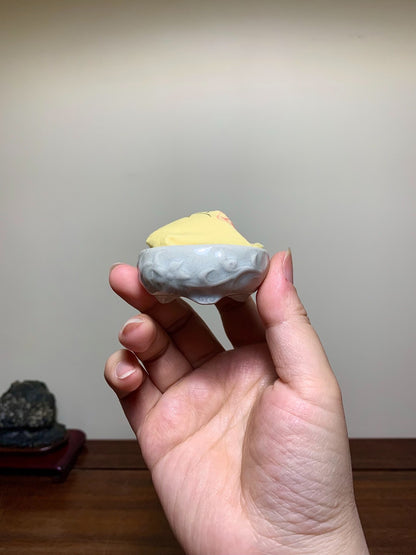 A hand holds a small, grey, glossy ceramic pot with a yellow lid and red mark.