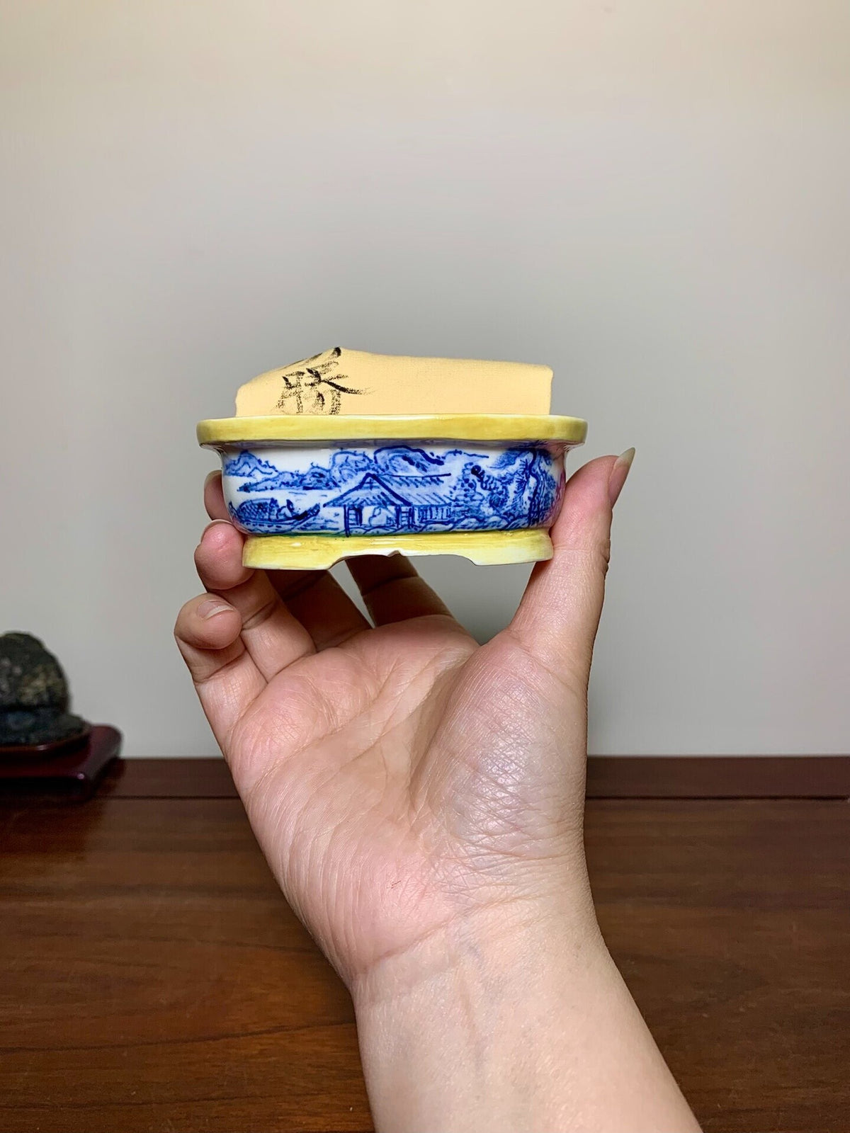 A hand holds a small blue and white ceramic pot with a yellow glazed lid featuring black brushwork.