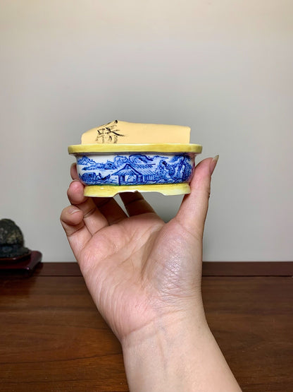 A hand holds a small blue and white ceramic pot with a yellow glazed lid featuring black brushwork.