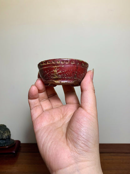A hand holds a small, deep red glazed pot with gold speckles and a row of raised dots around the rim.