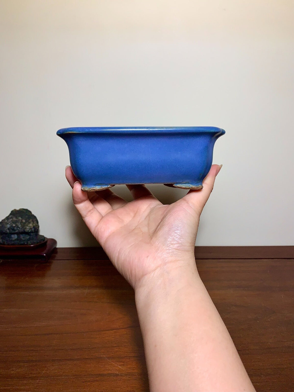 A hand holds a glossy, deep blue rectangular ceramic pot with slightly curved sides and unglazed foot rim.