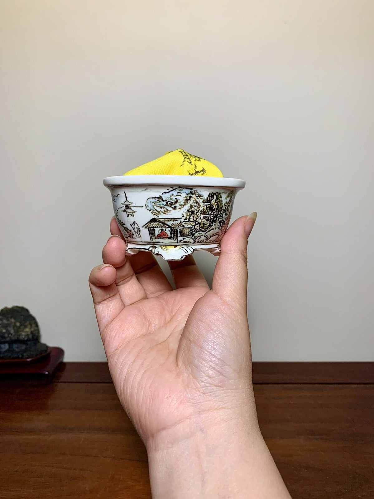 A hand holds a small white ceramic pot with black landscape painting and a yellow cloth inside.