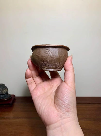 A hand holds a small, brown, unglazed ceramic pot with a carved bird and branch design.