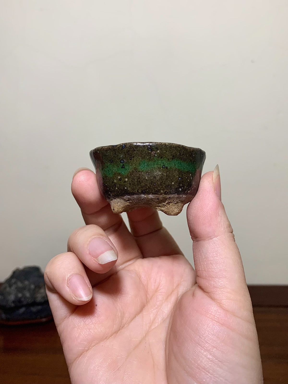 A hand holds a small, dark glazed pot with a green horizontal band and three feet.