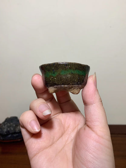 A hand holds a small, dark glazed pot with a green horizontal band and three feet.