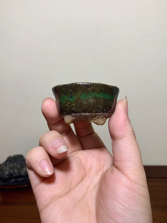 A hand holds a small, dark glazed pot with a green horizontal band and three feet.