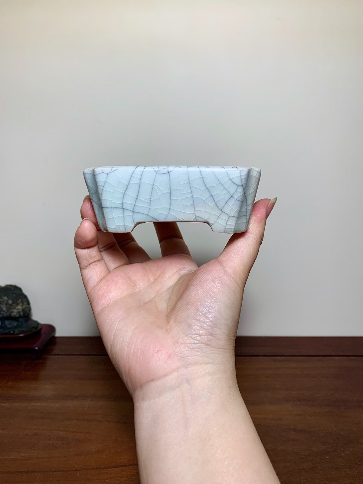 A hand holds a small, light grey, rectangular pot with a cracked glaze surface.