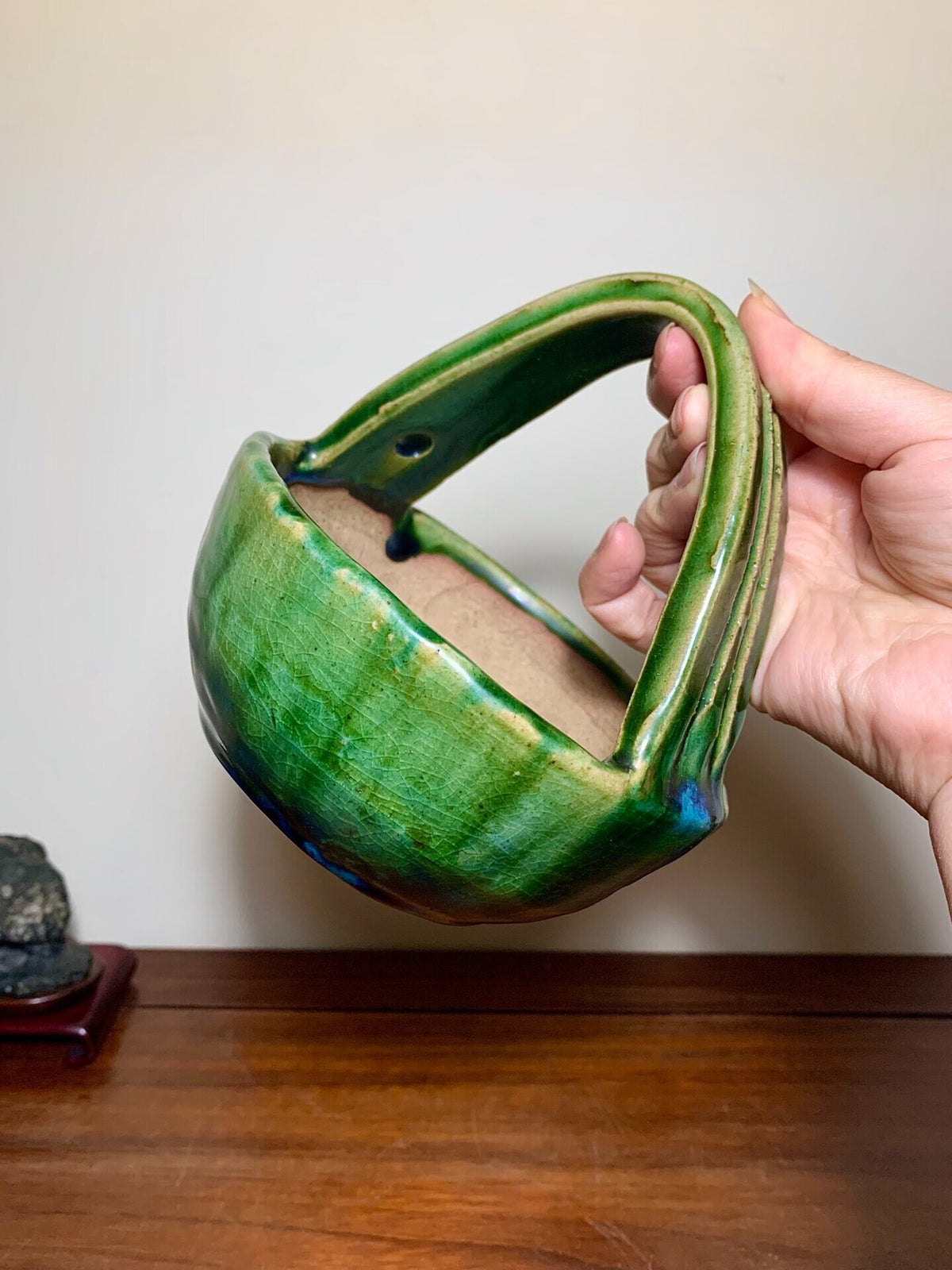 A hand holds a glossy green glazed ceramic pot with a curved handle and a small hole.