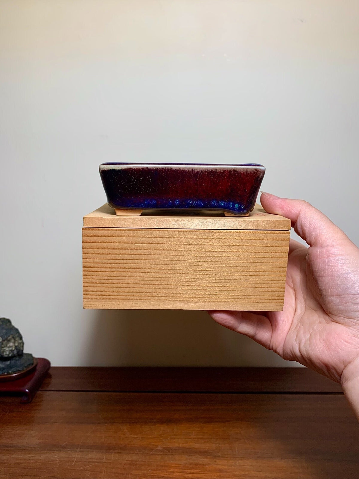 A hand holds a rectangular, glossy dark blue glazed pot on a light wood block.