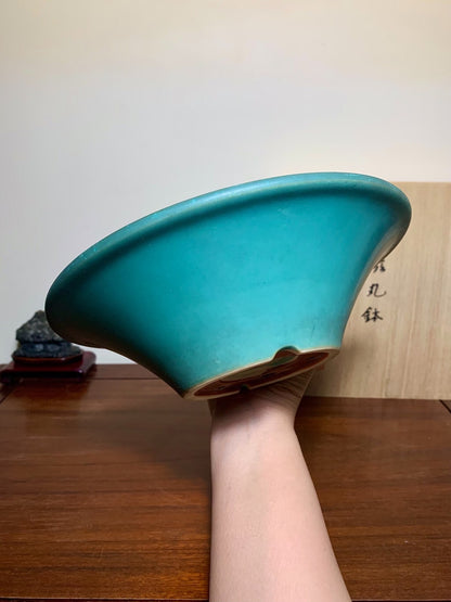 A hand holds a glossy teal ceramic pot with a flared rim and unglazed brown base.