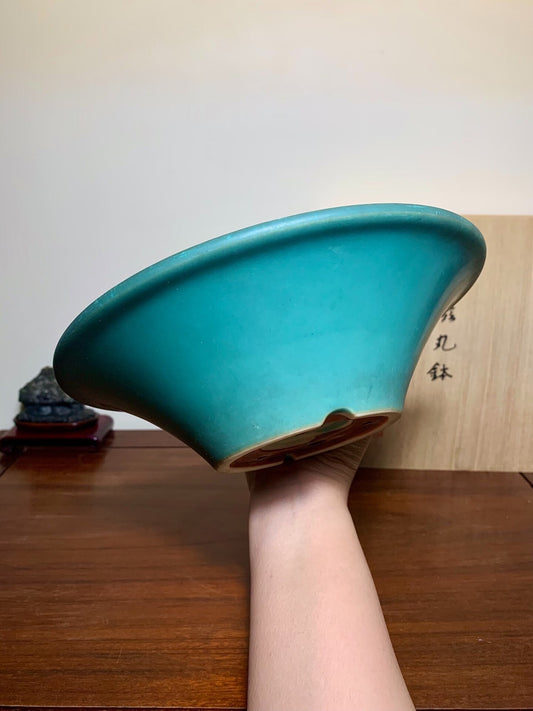 A hand holds a glossy teal ceramic pot with a flared rim and unglazed brown base.
