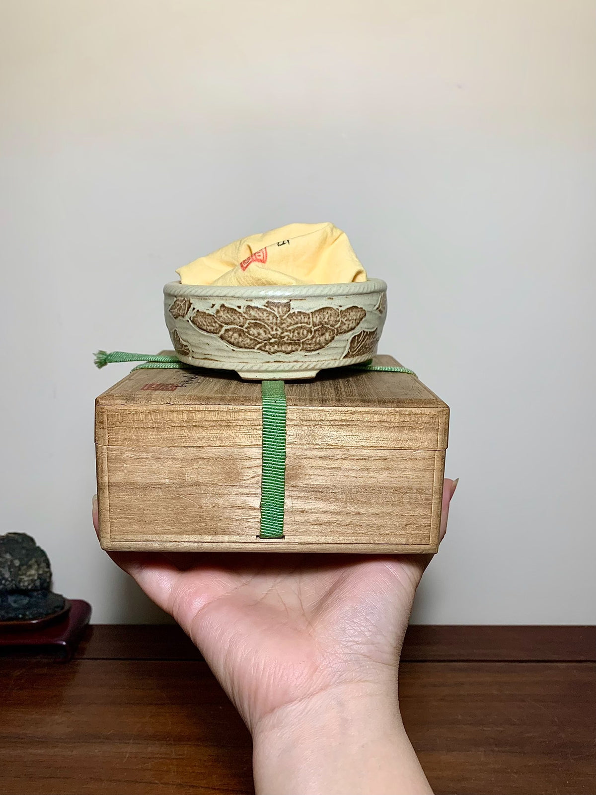 A hand holds a wooden box with a small, beige glazed pot featuring brown moth motifs.