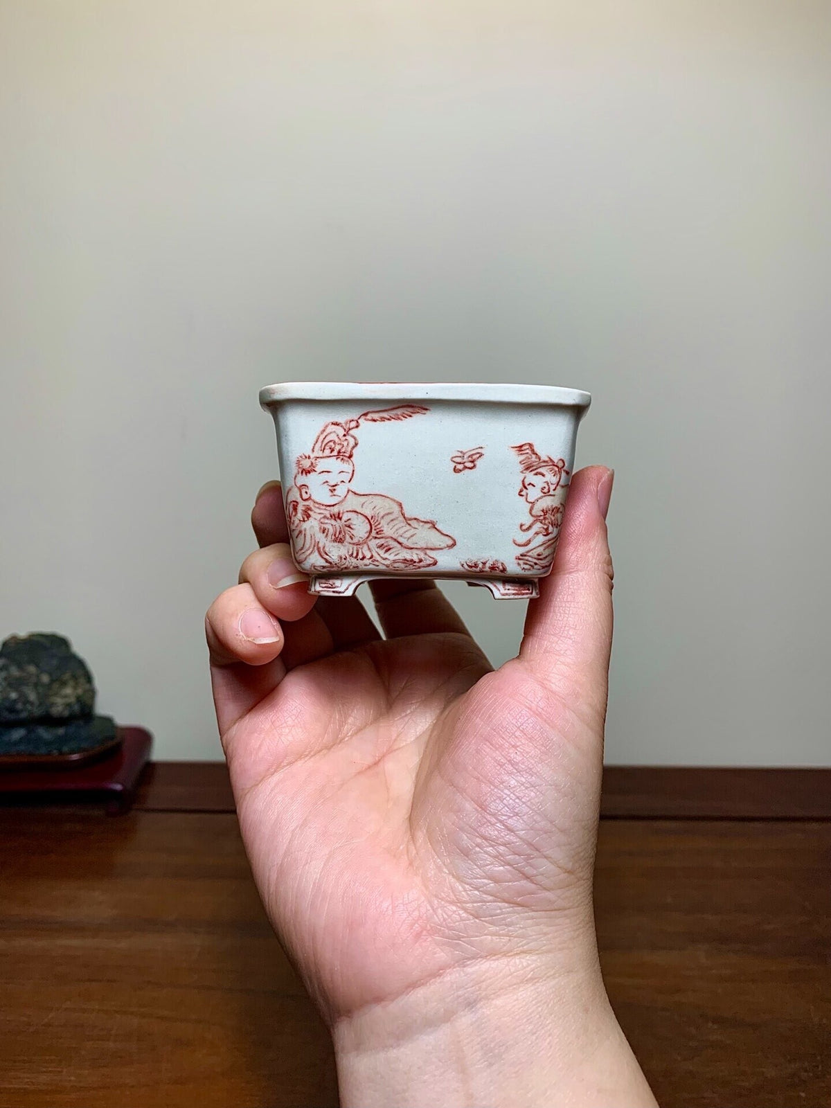 A hand holds a small, light grey glazed pot with red painted figures and a rabbit.