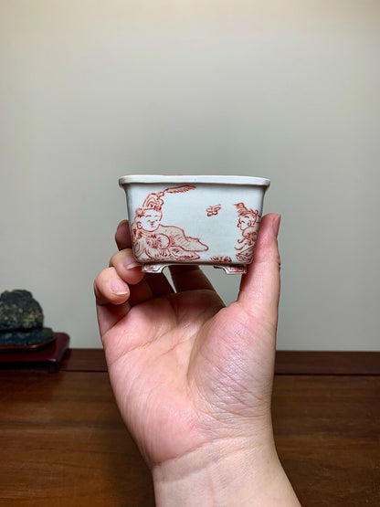 A hand holds a small, light grey glazed pot with red painted figures and a rabbit.