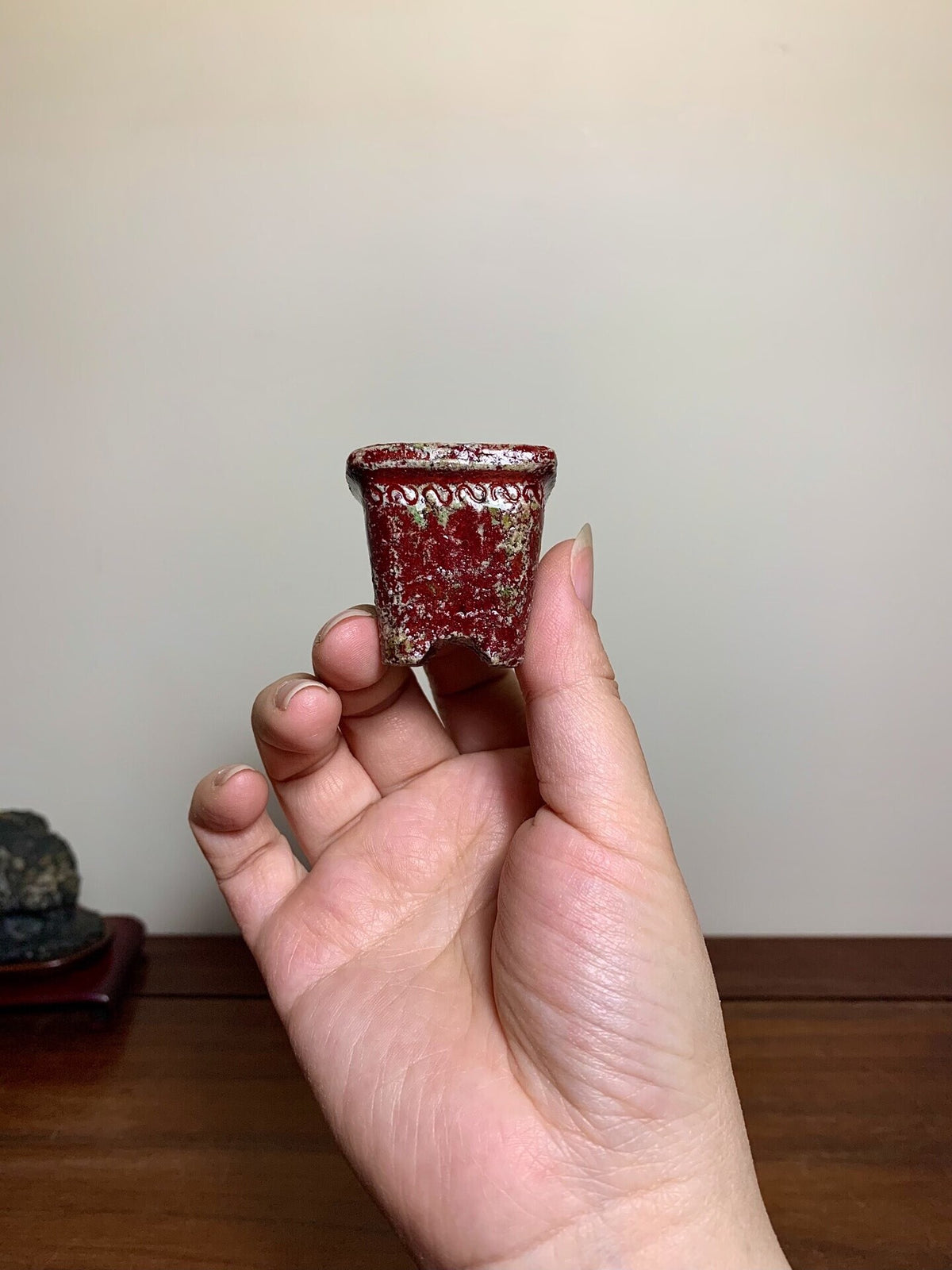 A hand holds a small, red glazed pot with white speckles and a decorative rim.