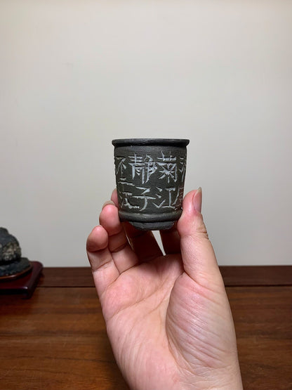 A small, dark grey, unglazed ceramic pot with white Japanese characters on its side.