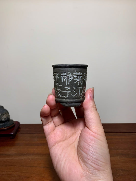 A small, dark grey, unglazed ceramic pot with white Japanese characters on its side.