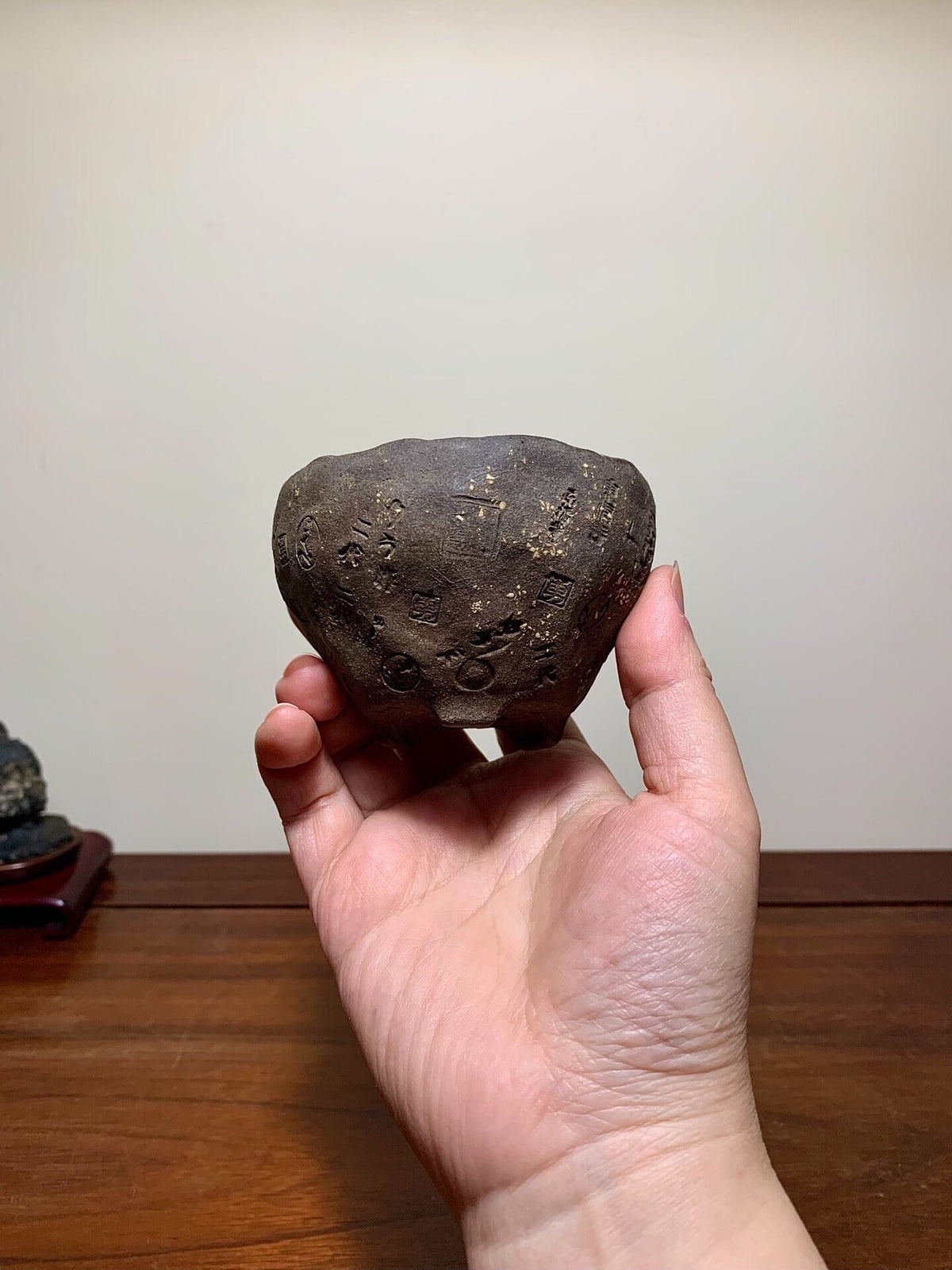 A hand holds a small, dark, unglazed pot with carved characters and a rough, textured surface.