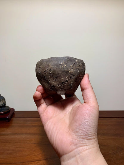 A hand holds a small, dark, unglazed pot with carved characters and a rough, textured surface.