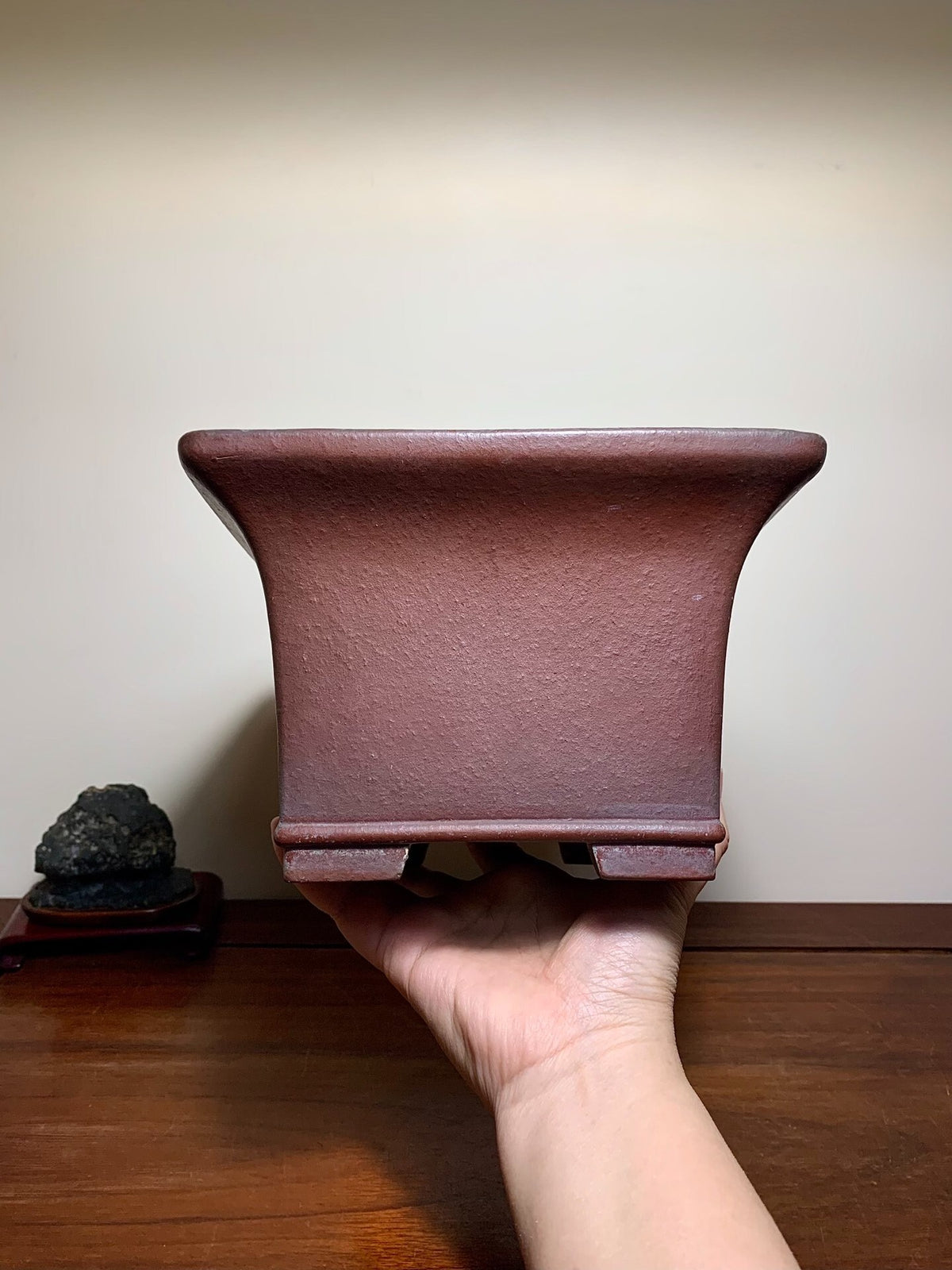 A hand holds a matte, reddish-brown, square bonsai pot with flared sides and unglazed surface.