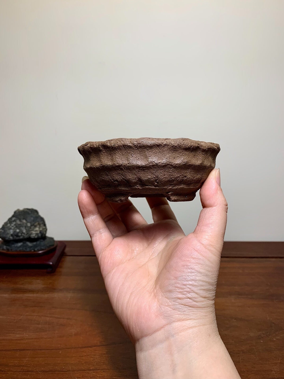A hand holds a small, brown, unglazed ceramic pot with textured, ridged sides.