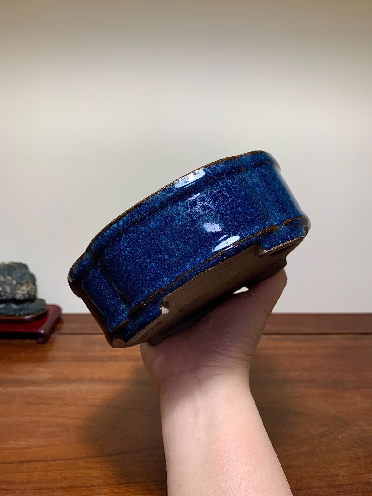 A hand holds a glossy, deep blue ceramic pot with a cracked glaze and brown rim.