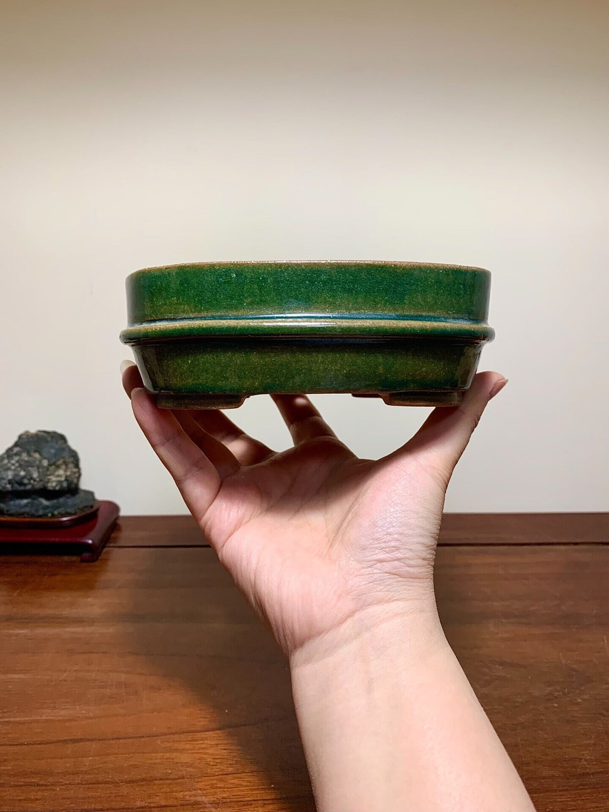 A hand holds a small, rectangular green glazed pot with a slightly raised rim.