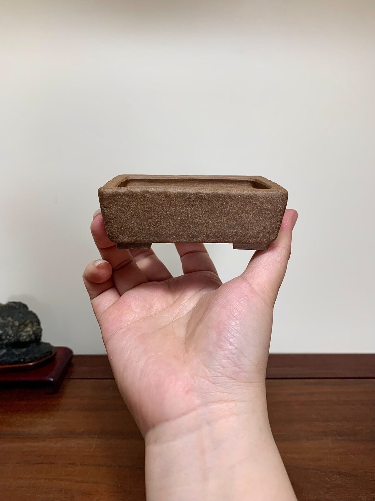 A hand holds a small, rectangular, unglazed brown ceramic pot with a matte finish.