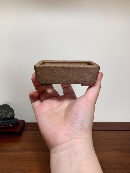 A hand holds a small, rectangular, unglazed brown ceramic pot with a matte finish.