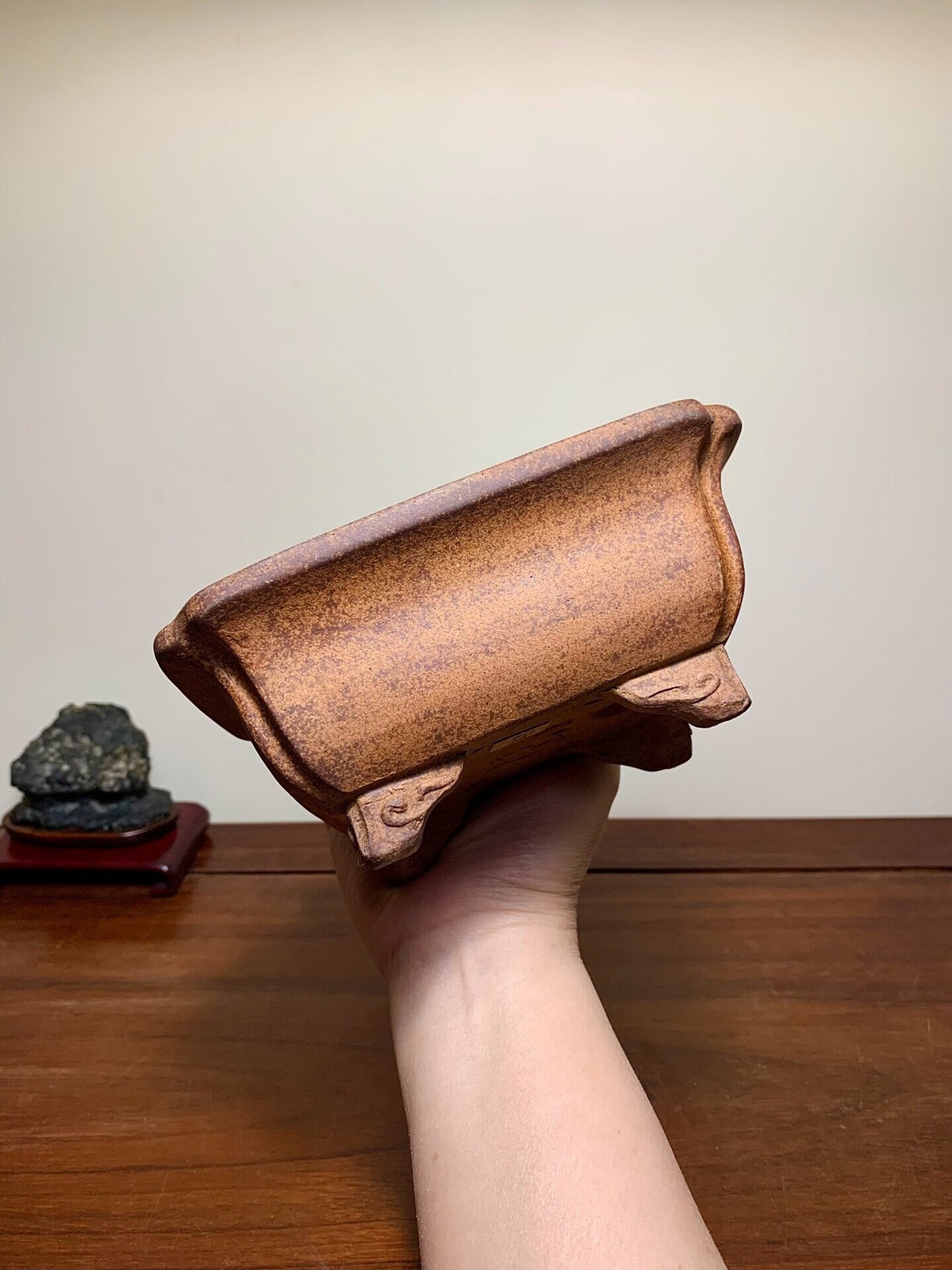 A hand holds a small, rectangular, unglazed brown ceramic pot with carved feet and a speckled surface.