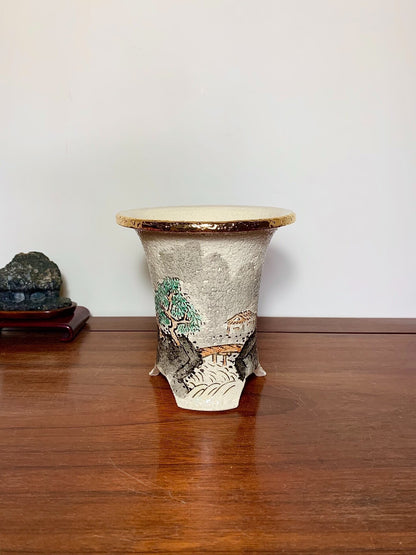 A grey glazed pot with a gold rim, featuring a painted landscape scene.
