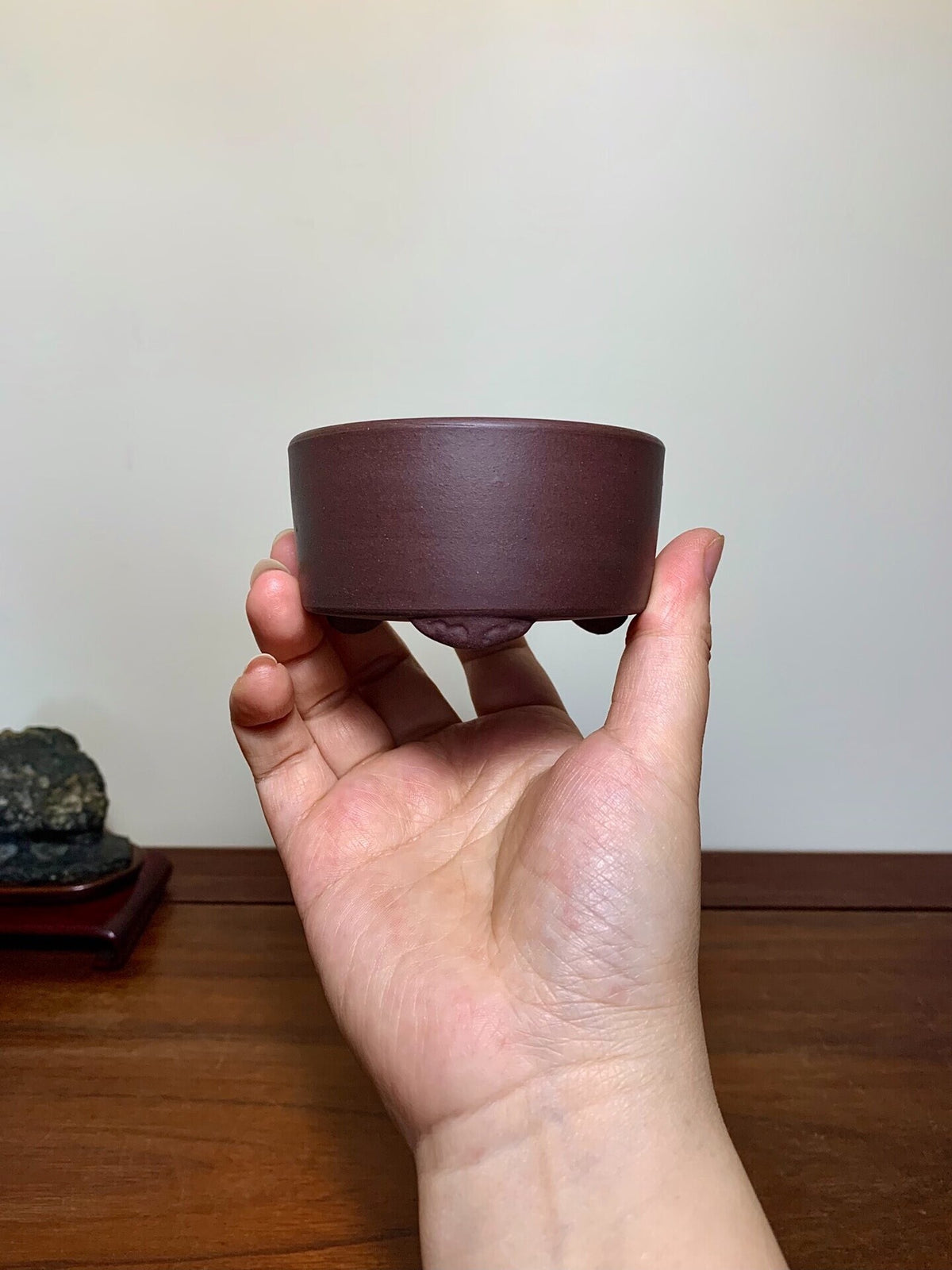 A hand holds a small, dark brown, matte-finished ceramic pot with a wide rim and three short feet.