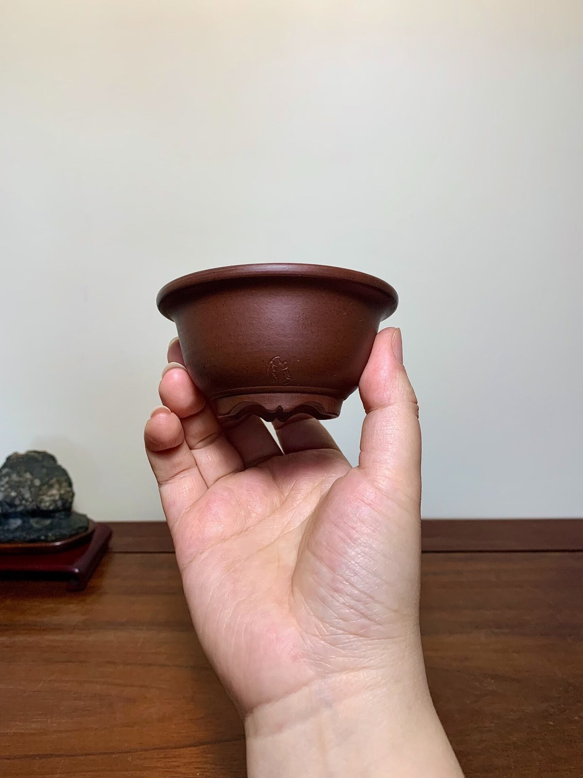A hand holds a small, dark brown, unglazed ceramic pot with a subtle impressed mark.