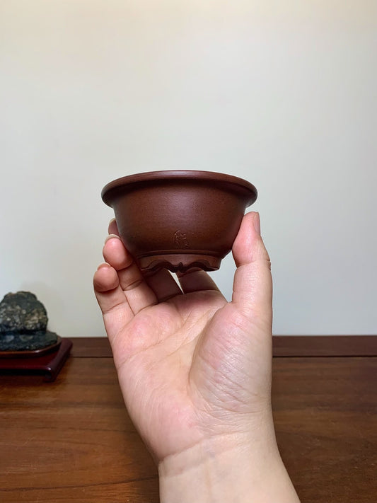 A hand holds a small, dark brown, unglazed ceramic pot with a subtle impressed mark.