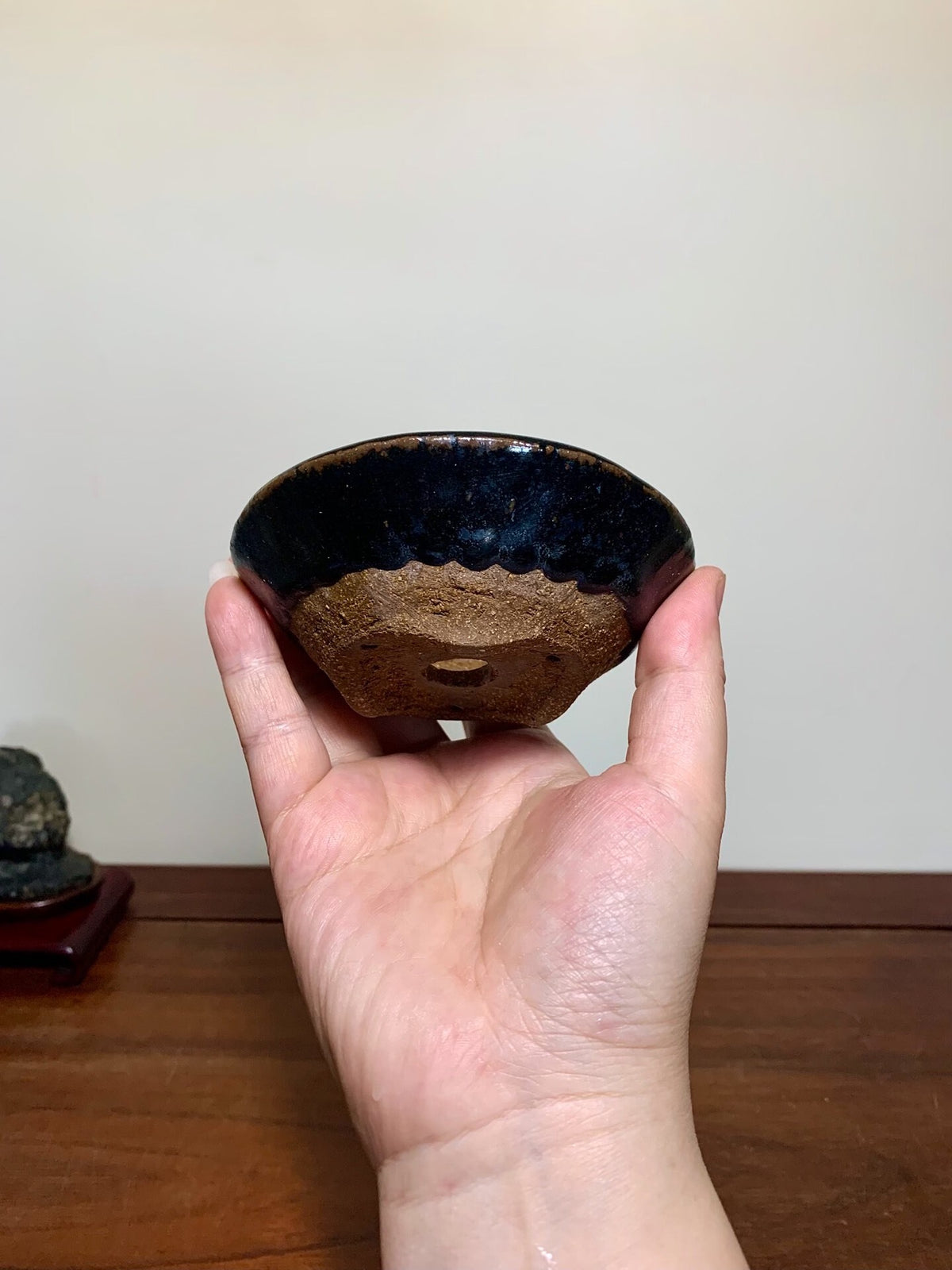 A hand holds a small, glossy black bowl with a rough, unglazed brown base rim.