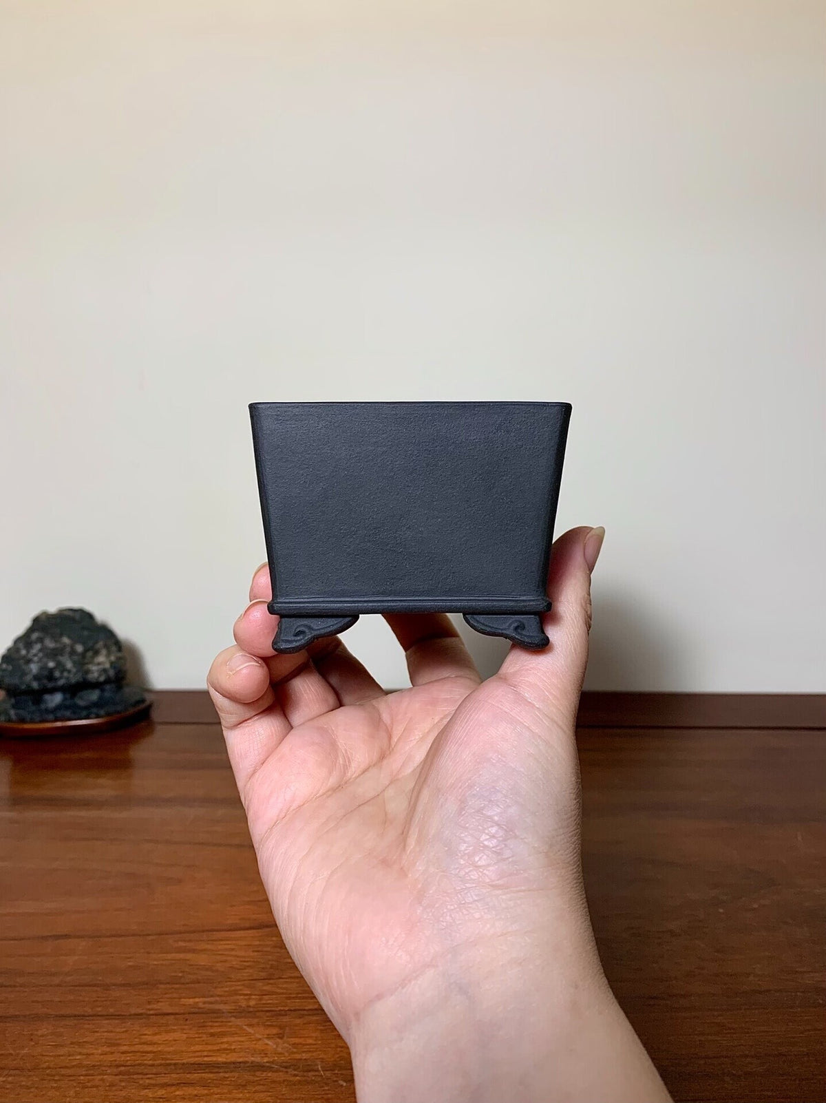 A hand holds a small, matte black square pot with four short feet.