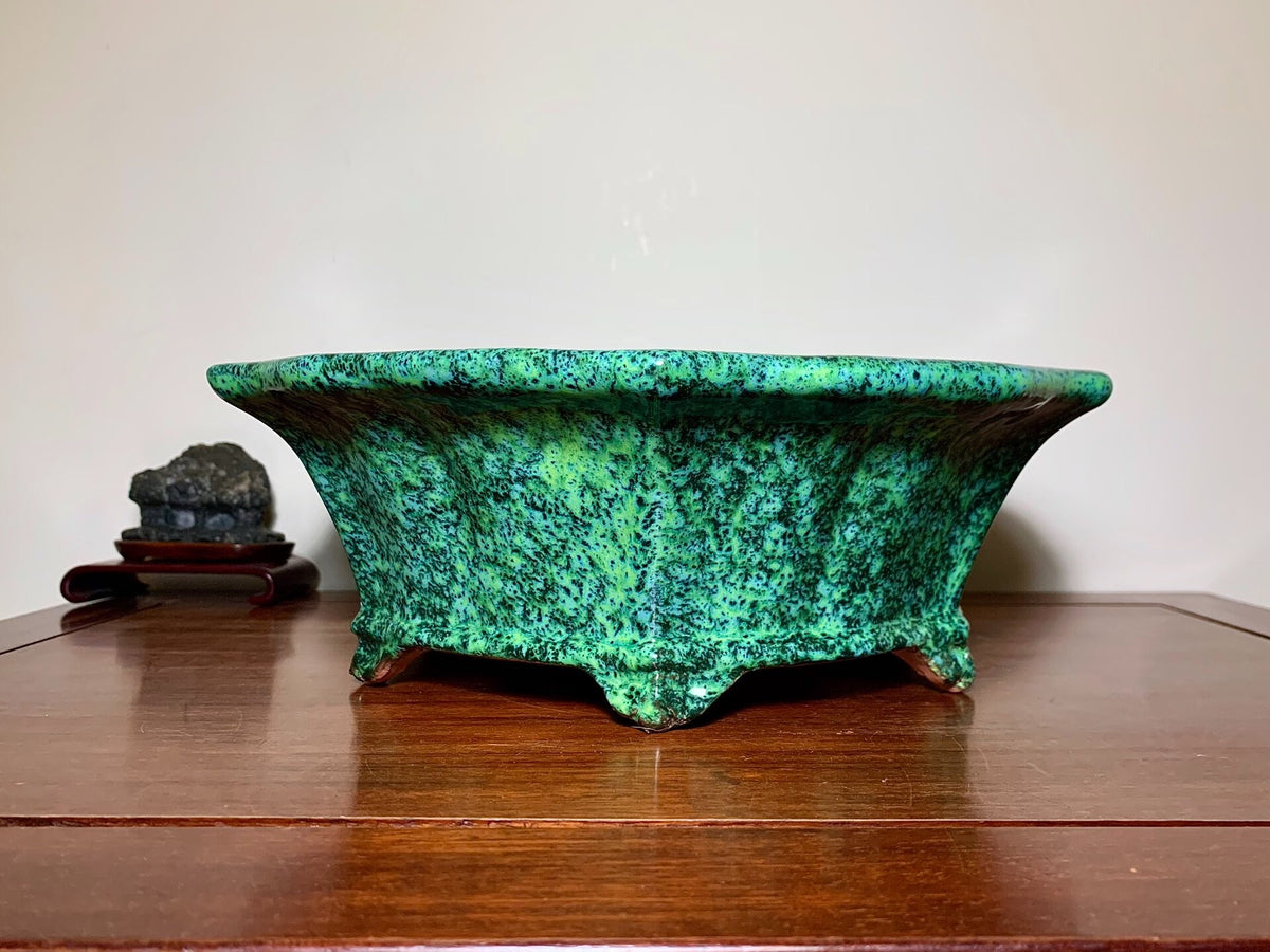 A wide, shallow green glazed pot with speckled texture and three feet, sits on a wooden surface.