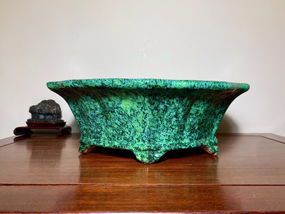 A wide, shallow green glazed pot with speckled texture and three feet, sits on a wooden surface.